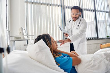 smart indian doctor talking to his african american female patient lying in bed in ward, healthcare clipart