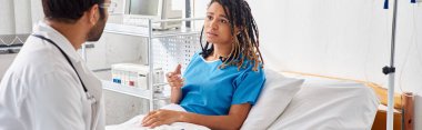 attractive ill african american woman lying in hospital bed talking to young indian doctor, banner clipart