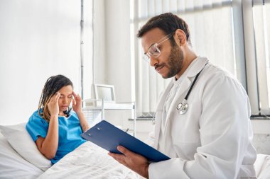 Baş ağrısı olan Afrikalı Amerikalı hastasının yanında yatağında oturan Hintli genç doktora odaklan.