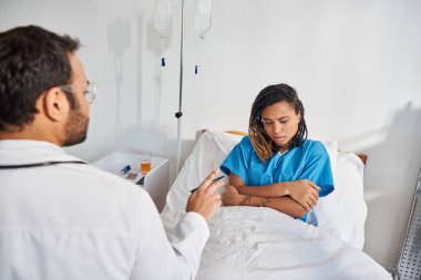 Çekici, hasta, Afro-Amerikalı bir kadın hastane yatağında yatıp Hintli genç doktorunu dinliyor.