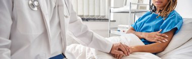 Afro-Amerikan bir hastanın elini tutan Hintli doktor hastane yatağında yatıyor.