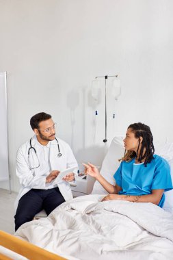 Afro-Amerikan hasta, hastane yatağında yatıp Hintli doktoruyla konuşuyor ve tablete not alıyor.