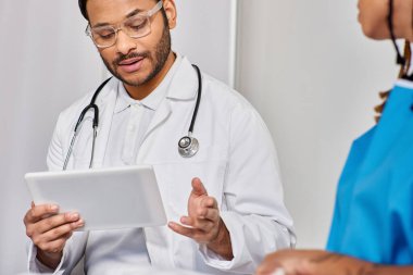 Afro-Amerikan kadının hastane yatağında tabletli bulanık Hintli doktora bakışı.