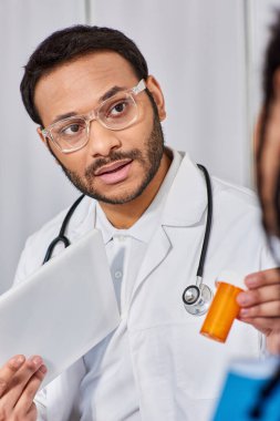 focus on indian doctor in glasses showing pills to his african american patient holding tablet clipart