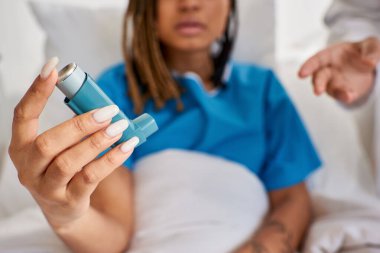 focus on asthma inhaler in hands of african american patient with doctor next to her in ward clipart