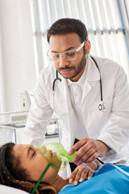 Genç Hintli doktor, hasta Afrikalı Amerikalı kadın hastasına oksijen maskeli, sağlık hizmetlerinde yardım ediyor.