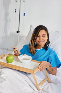 Afro-Amerikan kadın hastane koğuşunda kahvaltıda yulaf lapası ve elma yiyor.