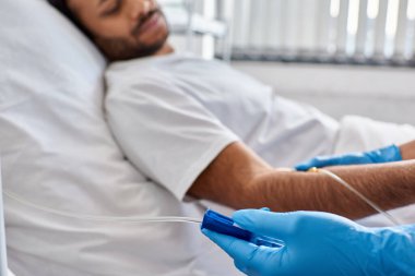 focus on hand of african american nurse setting drop counter with indian male patient on backdrop clipart