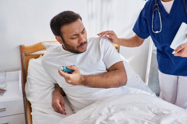 african american nurse helping her indian patient with asthma inhaler in his hospital ward clipart