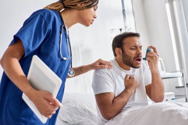 african american nurse helping her indian patient with asthma inhaler in his hospital ward clipart