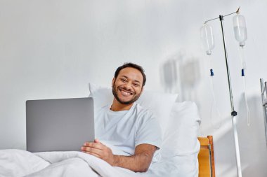 Hastane yatağında dizüstü bilgisayarla çalışırken kameraya gülümseyen neşeli Hintli genç adam.