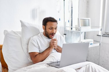 surprised jolly indian patient lying in hospital bed in his ward looking at his laptop, healthcare clipart