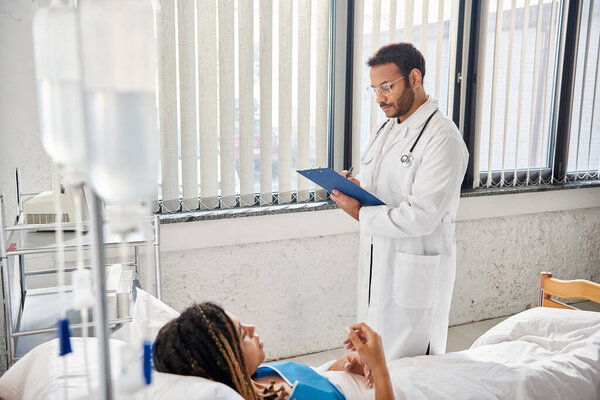 good looking indian doctor taking notes on african american patient lying in bed in hospital ward