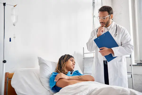 stock image pensive indian doctor looking at african american patient lying in bed in hospital ward, healthcare