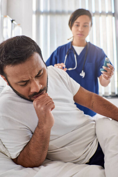 young african american nurse giving asthma inhaler to her ill indian patient in his ward, healthcare