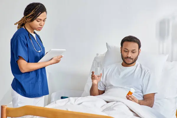 young african american nurse looking at tablet next to her indian patient holding pills, healthcare