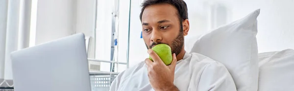 young indian man having video call and holding green apple while lying in hospital bed, banner