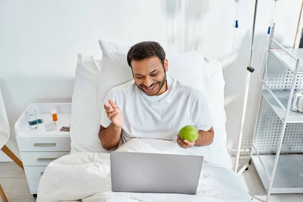 young indian man having video call and holding green apple while lying in hospital bed, healthcare