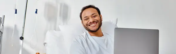 young cheerful indian man smiling at camera while working on laptop in hospital bed, banner