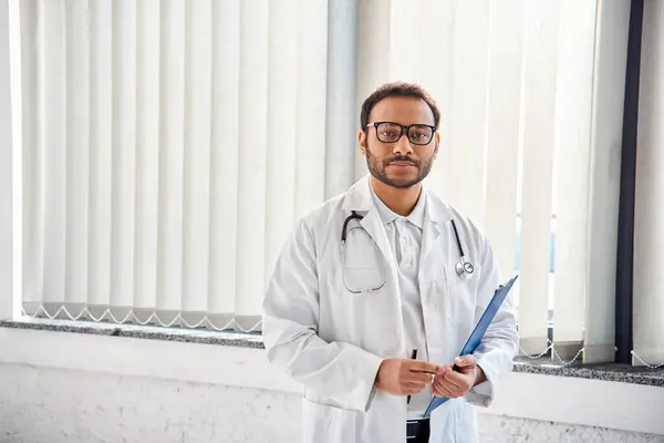 young indian doctor with glasses and stethoscope in hospital ward looking at camera, healthcare