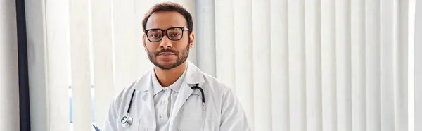 indian doctor with glasses and stethoscope in hospital ward looking at camera, healthcare, banner