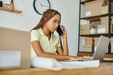 Güzel görünümlü Afro-Amerikan kadın bilgisayarında çalışırken telefonla konuşuyor, teslimat konsepti.
