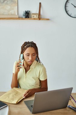 Güzel görünümlü Afro-Amerikan kadın elinde bir paketle telefonla konuşuyor ve dizüstü bilgisayarına bakıyor.