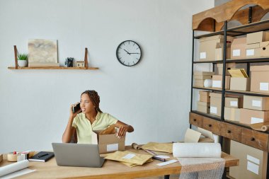 Neşeli, genç, Afro-Amerikan kadın satıcı paketleme kutusu telefonla konuşuyor ve başka tarafa bakıyor.