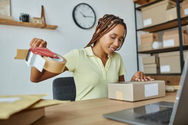 cheerful young african american female seller using tape to pack cardboard box and smiling happily clipart