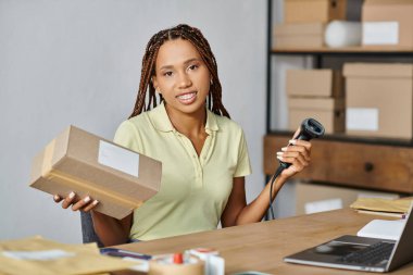 attractive jolly african american woman smiling at camera while working with scanner, delivery clipart