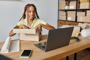 Neşeli genç Afro-Amerikan kadın tüccar paketleme kutusu ve dizüstü bilgisayara bakıyor, teslimat.