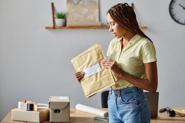 appealing young african american female seller looking at post packets during work, delivery concept clipart