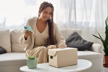 Güzel görünümlü Afro-Amerikan kadın elinde akıllı telefon ve karton pakete bakıyor.