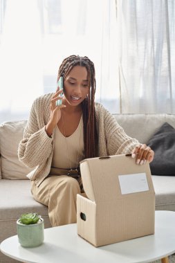 beautiful young african american woman in cozy homewear talking by phone while opening parcel clipart