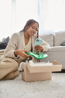 good looking joyous african american woman sitting on floor and taking photo of new green shoes clipart