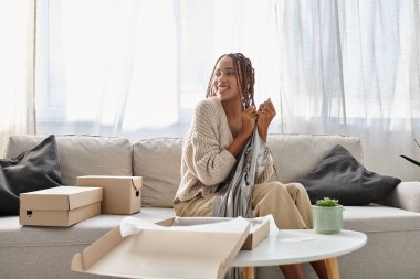 cheerful african american woman with braces unpacking her stylish silver gown while sitting on sofa clipart