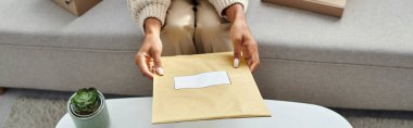 cropped view of young african american woman holding paper post packet above table, banner clipart