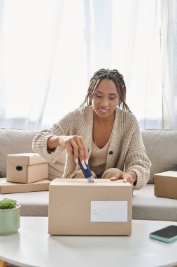 cheerful young african american woman sitting on sofa and using stationery knife to open parcel clipart