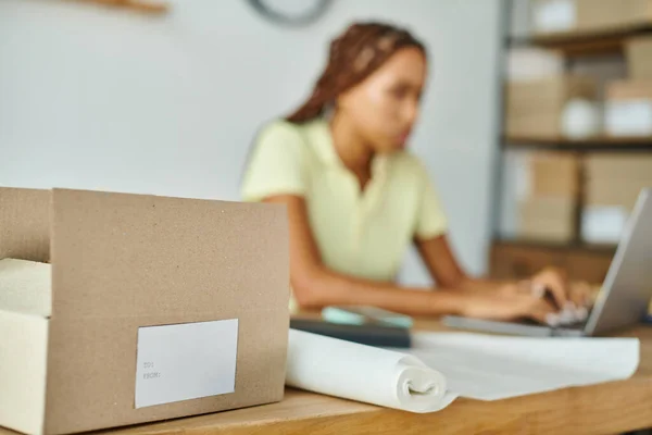 stock image focus on cardboard box with label with blurred african american female merchant on background