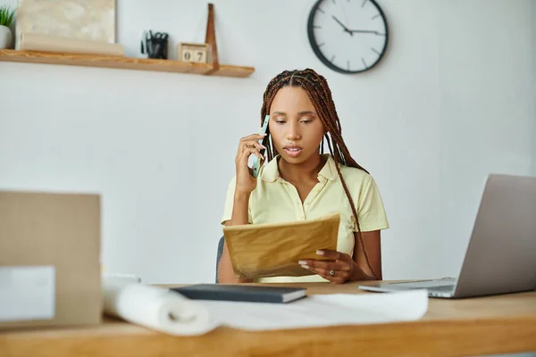 attractive african american female seller talking by phone and looking at post packet, delivery