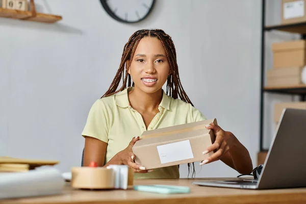 beautiful jolly african american female merchant holding box and looking at camera, delivery concept