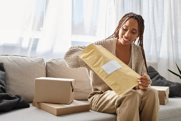 cheerful good looking african american woman opening post packet while sitting on couch at home