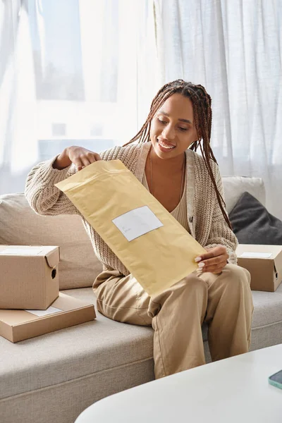 joyous african american woman in homewear opening post packets while sitting on sofa next to parcels
