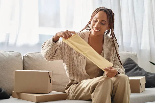 jolly young african american woman opening post packet while sitting on sofa surrounded by parcels