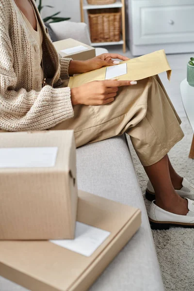 cropped view of young african american woman in homewear opening post packet while sitting on couch