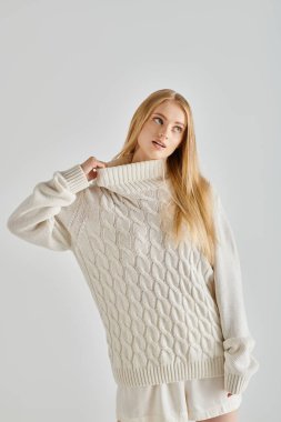 Düşünceli sarışın kadın. Gri arka plana bakarken sıcak kış süveteri yakasını esnetiyor.