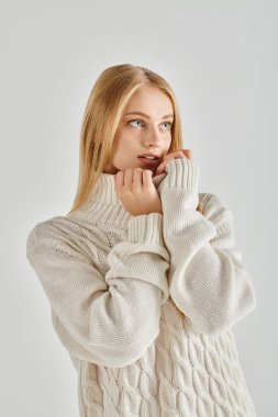 Beyaz kışlık süveter içinde büyüleyici ve hayalperest bir kadın model gri üzerinde dururken gözlerini kaçırıyor.