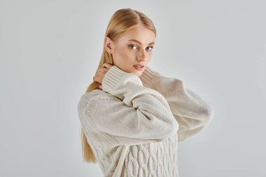 alluring blonde woman in white knitted sweater embracing own neck and looking at camera on grey clipart