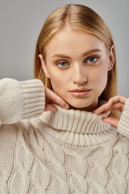 portrait of blonde woman with natural makeup and expressive gaze posing in knitted sweater on grey clipart