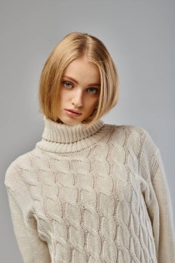 Beyaz örgü süveter giyen, kendine güvenen ve çekici sarışın kadın gri, soğuk mevsim kıyafetlerinde poz veriyor.